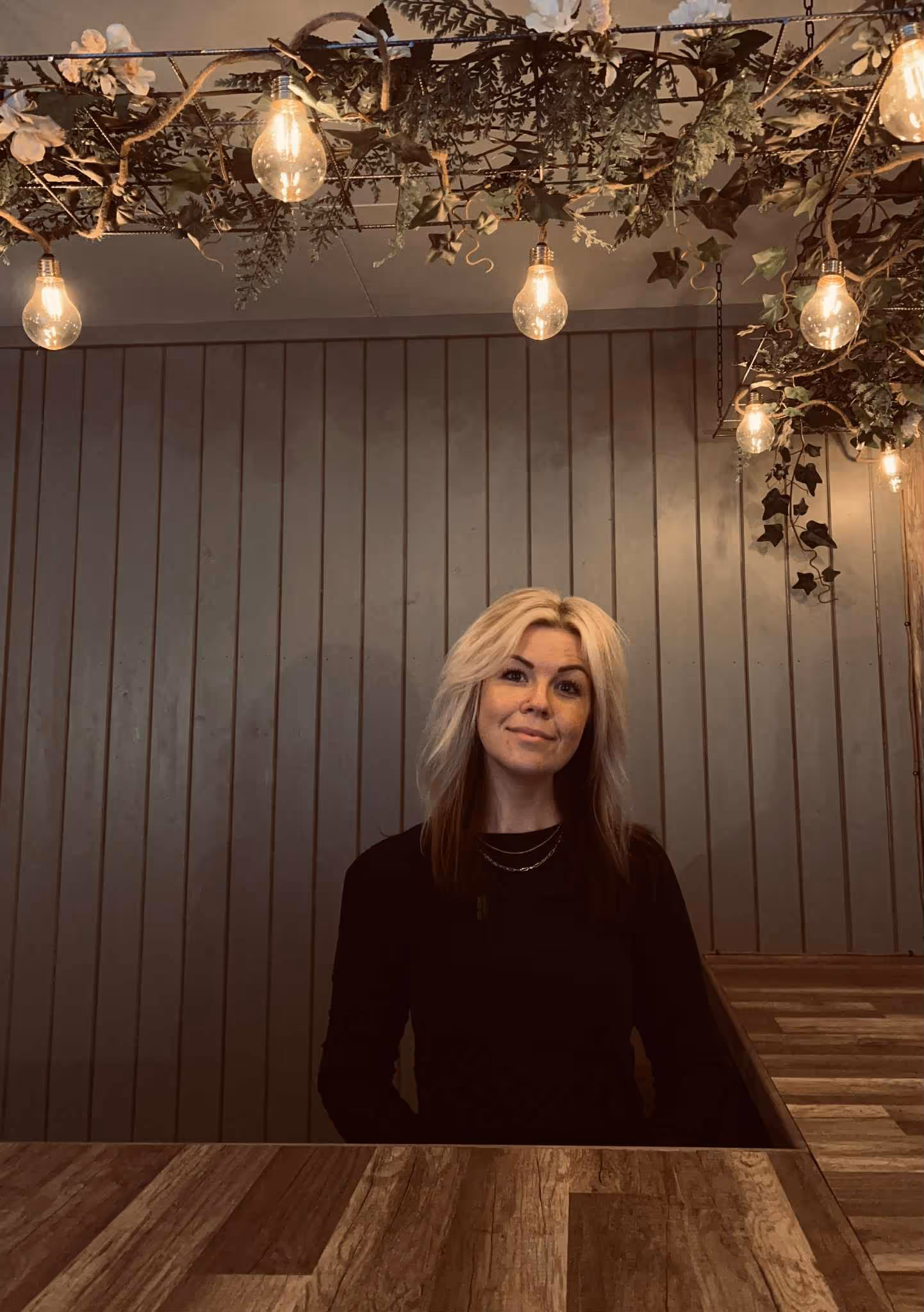 En kvinne med blondt hår sitter smilende ved en tredisk, under dekorative hengelamper og grønne planter mot en bakgrunn av vertikale trepaneler.